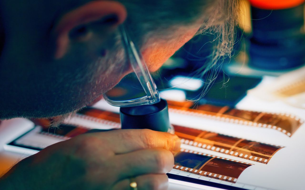 our-mission-01 A photographer closely inspects film negatives using a loupe and lightbox in a creative studio setting.